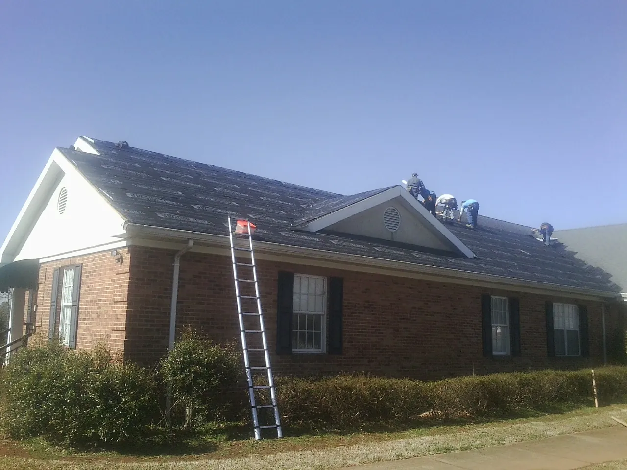 Crew tearing shingles off a brick ranch roof with a ladder set — Emergency Roof Repair work by our crew in Oxford Junction