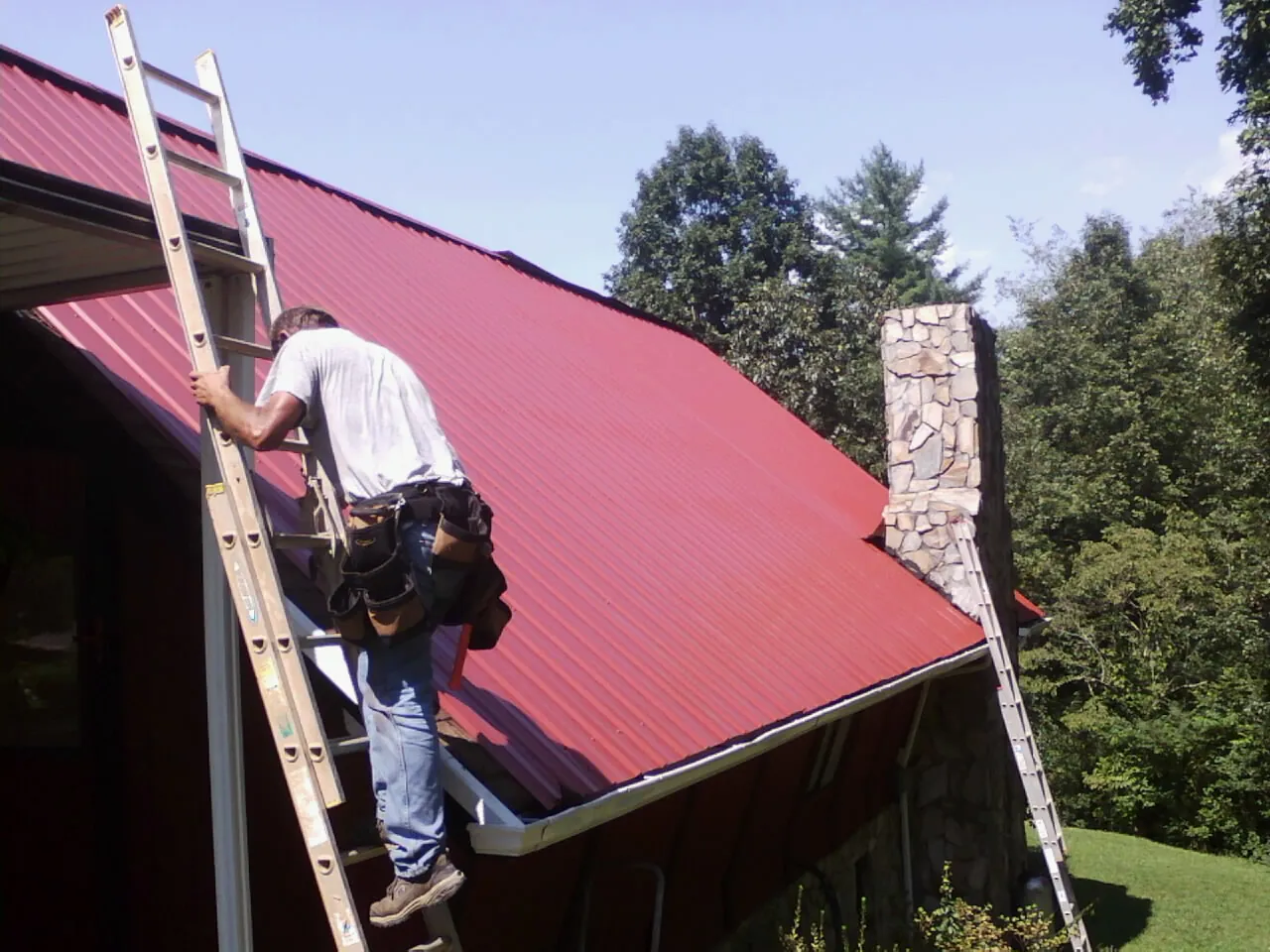 Roofer on a ladder beside a red metal roof and stone chimney — Soffit & Fascia Repair by Roofing Brothers in Oxford Junction